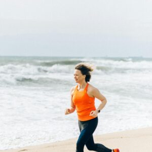 Caucasian woman jogging on the beach in orange tank top, embodying active lifestyle and fitness.